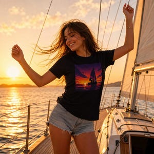 Puede incluir: Camiseta negra con un gráfico de velero al atardecer. La imagen muestra a una persona en un velero con los brazos levantados, usando pantalones cortos de mezclilla. El fondo es una puesta de sol sobre el océano, con la vela del barco visible.
