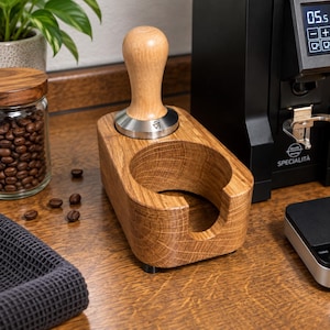 Könnte beinhalten: Eine Kaffeetamper-Station aus hellbraunem Holz mit einer kreisförmigen Aussparung und einem passenden Tamper mit Holzgriff und Edelstahlbasis. Die Station weist eine sichtbare Holzmaserung auf und steht auf einer flachen Oberfläche.