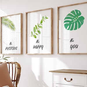 Könnte beinhalten: Drei gerahmte Drucke mit grünen Blättern und den Worten "Be Positive", "Be Happy" und "Be You" hängen an einer weißen Wand über einer weißen Kommode mit brauner Oberseite und einem Korbstuhl mit brauner Wolldecke.