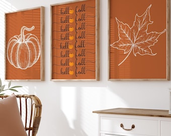 May include: Three framed prints with fall-themed designs. The left print features a white outline of a pumpkin on an orange background. The center print has the text "hell fall" repeated eight times in black on an orange background. The right print features a white outline of a maple leaf on an orange background.