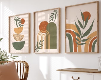 May include: Three abstract art prints with a boho theme featuring geometric shapes, leaves, and a rainbow design in shades of green, orange, and brown. The prints are framed in natural wood and hung on a white wall above a white cabinet with a wooden top.