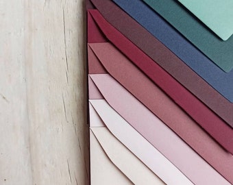 May include: A set of envelopes in various shades of green, blue, burgundy, pink, and beige. The envelopes are arranged in a fan shape, with the darkest color at the top and the lightest color at the bottom.