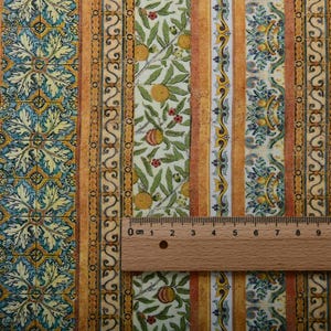 May include: Fabric with a repeating pattern of vertical stripes. The design includes floral and fruit motifs, with oranges and leaves. The color palette features shades of yellow, orange, green, and blue. A wooden ruler is placed on the fabric, showing measurements in centimeters.