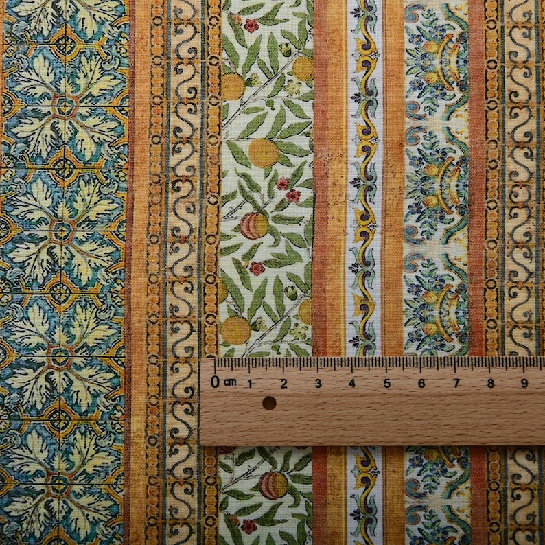 Op de afbeelding: Stof met een herhalend patroon van verticale strepen. Het ontwerp bevat bloemen- en fruitmotieven, met sinaasappels en bladeren. Het kleurenpalet bevat tinten geel, oranje, groen en blauw. Een houten liniaal ligt op de stof en toont de afmetingen in centimeters.
