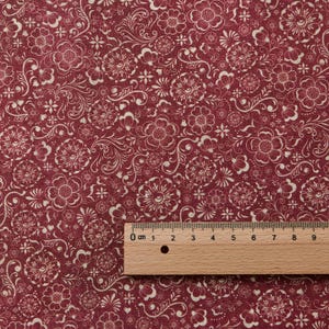 Op de afbeelding: Bordeaux stof met een crèmekleurig bloemenpatroon. Het ontwerp omvat gestileerde bloemen, bladeren en krullen. Een houten liniaal met centimeter markeringen is op de stof geplaatst, met een meting van ongeveer 9 cm.