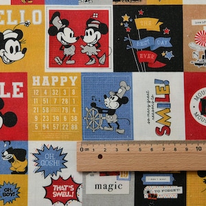 Könnte beinhalten: Ein farbenfroher Stoff mit verschiedenen Quadraten, die Illustrationen von Mickey Mouse und Minnie Mouse zeigen. Der Stoff enthält Texte wie "HAPPY", "SMILE!" und "THE BEST DAY EVER". Ein Holzlineal liegt auf dem Stoff und zeigt Messungen in Zentimetern an.