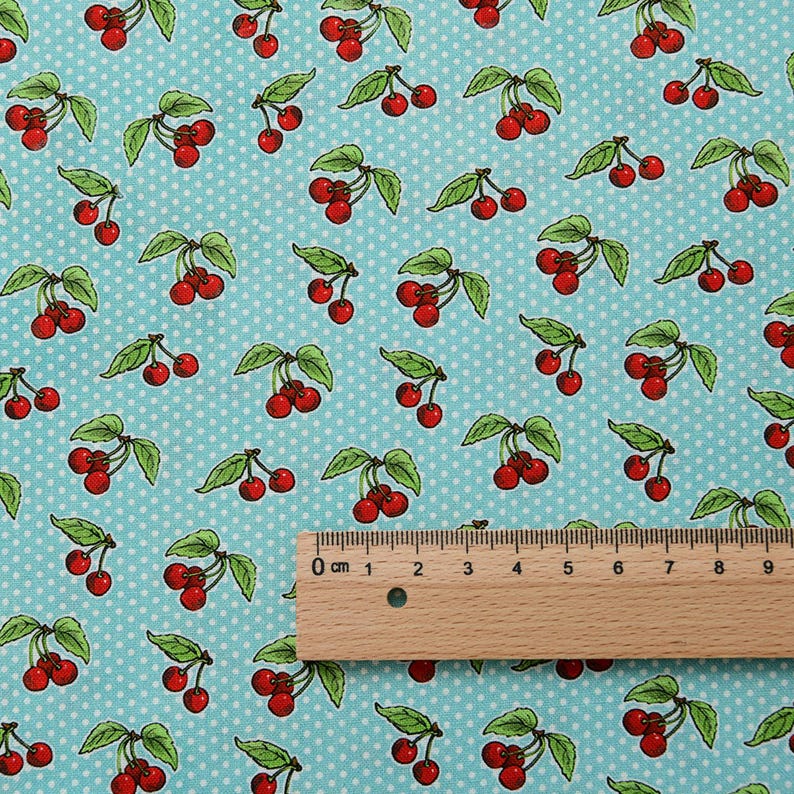 May include: Fabric featuring a repeating pattern of red cherries and green leaves on a light blue background with white polka dots. A wooden ruler is placed on the fabric, showing measurements in centimetres.
