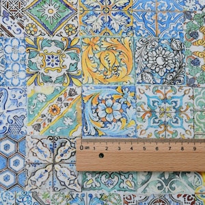 May include: Close-up of a fabric with a repeating pattern of colorful, square tiles. Each tile features a unique design with floral and geometric motifs in shades of blue, yellow, green, and white. A wooden ruler is placed across the tiles, showing centimeter measurements.