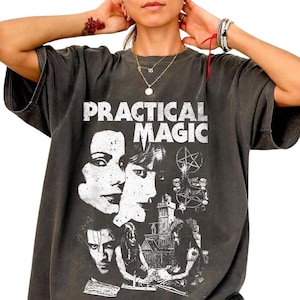 以下が含まれることがあります： 上部に「PRACTICAL MAGIC」の文字が白くプリントされた、チャコールグレーの半袖Tシャツ。デザインには、2人の人物の様式化されたポートレート、五芒星のシンボル、そして家の詳細なイラストが含まれています。