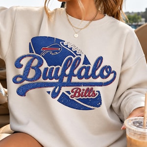 Puede incluir: Una sudadera beige de cuello redondo con un gran gráfico desgastado de un balón de fútbol americano azul y el texto Buffalo Bills en una fuente cursiva azul con bordes rojos. El diseño incluye un pequeño logotipo de un bisonte.