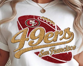 Può includere: Una t-shirt bianca a maniche corte con una grande grafica di un pallone da football americano rosso con una texture dorata usurata. La grafica include il testo 49ers in un grande carattere dorato e San Francisco in un corsivo dorato.