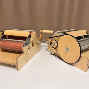 May include: Two wooden fiber carders with a light wood finish. Each carder has a red drum, a wooden handle, and a tray. The carders are designed for processing fibres.
