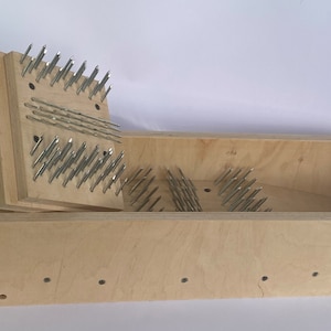 May include: A light-coloured wooden box with a removable lid, both with numerous metal pins. The box is rectangular, with the lid partially removed, revealing the interior. The pins are evenly spaced and appear to be securely fastened.