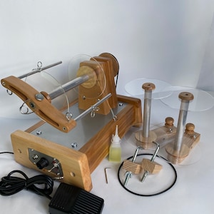 Può includere: Un avvolgitore di filato in legno con pedale, dischi in plastica trasparente e vari accessori. L'avvolgitore ha una finitura in legno naturale e una base in metallo argentato. Include una piccola bottiglia di olio e una chiave esagonale.