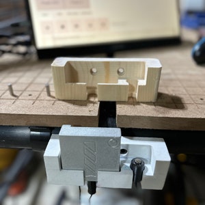 May include: A wooden and metal workshop tool is shown. The wooden piece is light-colored with two holes and a rectangular cutout. The metal piece is silver with a logo. The tool is mounted on a brown surface.