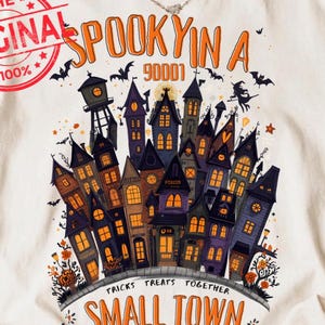 May include: An off-white short-sleeve t-shirt featuring a graphic of a cluster of dark purple and orange haunted houses. The text reads SPOOKY IN A SMALL TOWN with TRICKS TREATS TOGETHER and a red circular stamp that says THE ORIGINAL 100%.
