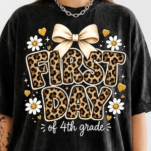 May include: A black mineral-washed t-shirt featuring a graphic with the text FIRST DAY of 4th grade. The letters have a brown and black leopard print pattern. The design includes a large cream-colored bow, white daisies, and small gold hearts.