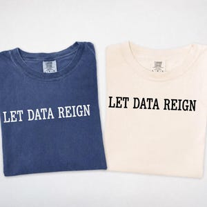 Puede incluir: Dos camisetas de cuello redondo, una en azul desteñido y otra en color crema. Ambas camisetas presentan el texto LET DATA REIGN impreso en una fuente negrita sin remates en el pecho. La camiseta azul tiene texto blanco y la crema texto negro.
