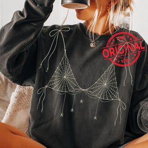 May include: A charcoal gray long-sleeve sweatshirt featuring a white graphic print of a bikini top made of spiderwebs with dangling spiders and tied strings. A red circular stamp with the text THE ORIGINAL is visible on the upper right side.