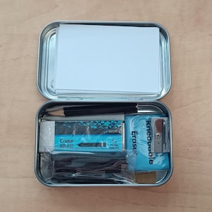 Könnte beinhalten: Eine rechteckige silberne Metalldose, die im Deckel einen Stapel weißer Papierkarten enthält. Der Boden enthält einen schwarzen Bleistift, einen weißen Radiergummi mit der Aufschrift Eraser RB-912, einen blauen Knetradierer, einen Anspitzer und schwarze Teile in Plastik.