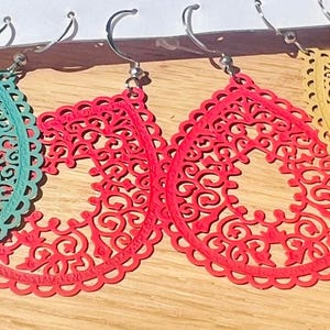 Könnte beinhalten: Türkis, rote und gelbe Tropfenohrringe mit filigranem Muster und silberfarbenen Haken. Die Ohrringe werden auf einer hellen Holzoberfläche präsentiert. Die Ohrringe haben ein offenes, spitzenartiges Design.