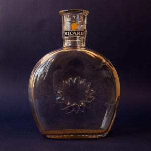 Peut inclure: Bouteille en verre transparent avec un corps arrondi et un col étroit. La bouteille présente un motif de tournesol gravé dans le verre et une étiquette avec le texte "RICARD" en noir et un logo soleil jaune. La bouteille est vide.