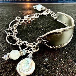 May include: A silver spoon bracelet with a decorative floral design on the spoon bowl. The bracelet features a chain with dangling charms, including a large pearl and several small, round, white beads. The bracelet is set against a dark background.
