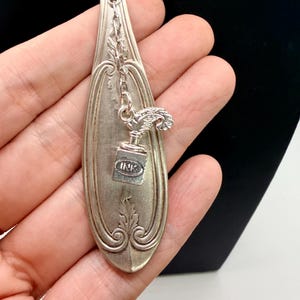 May include: A silver-toned pendant necklace featuring a repurposed spoon design. The spoon has ornate detailing and a small charm of an ink bottle and quill. The word "INK" is visible on the charm. The necklace is displayed against a black background.