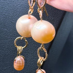 May include: A pair of gold-tone drop earrings featuring a teardrop-shaped clear stone at the top, a large round peach-colored pearl in the center, and a smaller oval peach-colored pearl dangling from a gold ring at the bottom of each earring.