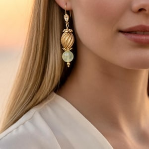 May include: A gold-toned drop earring featuring a small clear crystal at the top, a central textured gold-toned bead with a spiral design, and a smooth pale green spherical bead at the bottom, all suspended from a gold-toned hook wire.