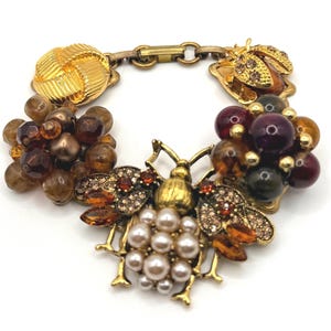 May include: A gold-toned bracelet featuring a large bee pendant with pearl accents and amber-colored crystals. The bracelet includes clusters of brown and purple beads and a decorative clasp.