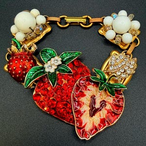 May include: A gold-tone link bracelet featuring strawberry charms decorated with red crystals and enamel, green enamel leaves, white flower accents with clear stones, and clusters of white beads and small clear crystals connected by a metal clasp.