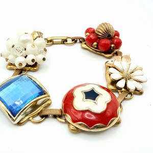 May include: A gold-tone charm bracelet featuring a variety of decorative elements including a cluster of white beads, a square blue faceted stone, a red and white floral-shaped charm with a dark center, a white daisy, and a cluster of red beads.
