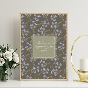 Puede incluir: Una impresión enmarcada con un fondo marrón claro y un patrón repetitivo de pequeñas flores azules. La impresión presenta un borde de encaje blanco alrededor de un rectángulo verde claro con el texto "En esta casa, hacemos risas, abrazos y gracia."