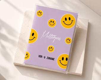 May include: A rectangular light purple notebook cover featuring a pattern of yellow smiley faces. The cover displays the white cursive text Mutterpass and the black printed text VOR- & ZUNAME. The notebook is enclosed in a clear plastic sleeve.