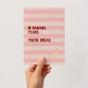 Könnte beinhalten: Eine Grußkarte mit rosa-weiß gestreiftem Hintergrund. Auf der Karte steht der Text "IN BANANA YEARS YOU'RE BREAD". Eine kleine Bananenillustration befindet sich unten rechts. Die Karte wird von einer Hand gehalten.