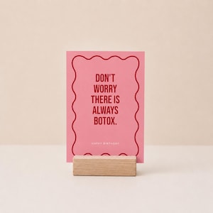 Könnte beinhalten: Eine rechteckige rosa Grußkarte mit gewelltem rotem Rand, die den roten Text DON'T WORRY THERE IS ALWAYS BOTOX. und HAPPY BIRTHDAY in kleineren weißen Buchstaben zeigt, aufrecht gehalten von einem rechteckigen hellen Holzblockständer.