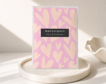 May include: A rectangular protective cover for a Mutterpass featuring a repeating pattern of light peach hearts on a pink background. A black horizontal rectangular label is centered on the front with the white text Mutterpass - Vor- & Zuname -.