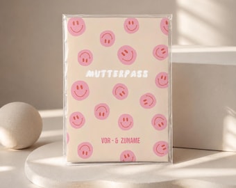 Puede incluir: Un folleto rectangular con una cubierta de color beige claro que presenta un patrón repetitivo de caras sonrientes rosas. El texto MUTTERPASS está impreso en blanco en el centro, y el texto VOR- & ZUNAME está impreso en rosa cerca de la parte inferior.