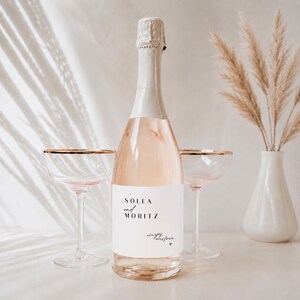 Könnte beinhalten: Eine Flasche Rosé-Sekt mit einem weißen Etikett, das den Text Solea und MORITZ sowie einen kleinen handschriftlichen Schriftzug trägt. Die Flasche wird von zwei klaren Glaskelchen mit Goldrand auf einem neutralen, hellen Hintergrund flankiert.