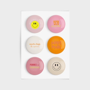 Könnte beinhalten: Sechs runde Knöpfe in Rosa, Orange und Creme mit verschiedenen Sprüchen und Smiley-Gesichtern. Die Sprüche lauten "Born to be Wild", "Snacks, hugs & Aperol", "Wmn Pwr" und "Pommes & Champagner".
