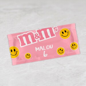 Könnte beinhalten: Rosa M&M's Süßigkeitenbeutel mit dem Text "MALOU 6" und mehreren gelben Smiley-Gesichtern.