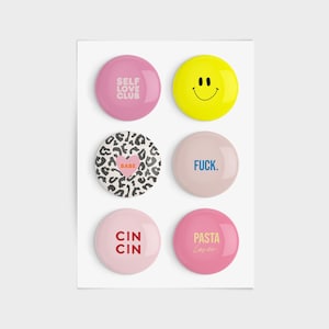 Könnte beinhalten: Sechs runde rosa, beige und gelbe Knöpfe mit verschiedenen Sprüchen und Designs. Die Knöpfe zeigen die Sätze "Self Love Club", "Babe", "F.U.C.K.", "Cin Cin", "Pasta Love" und ein Smiley.