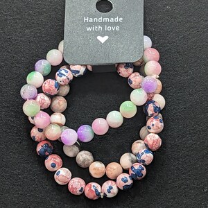 Op de afbeelding: Drie gelaagde armbanden gemaakt van roze, witte en groene kralen met bloemmotieven. De armbanden liggen op een zwarte achtergrond met een label waarop "Handmade with love" staat.