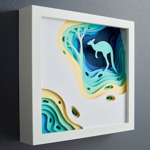 Puede incluir: Una pieza de arte de papel enmarcada en blanco con un diseño en capas de papel azul, verde y amarillo. El diseño presenta una silueta de un canguro de pie en una colina.