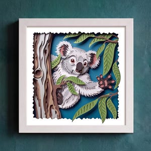 Puede incluir: Una obra de arte en papel multicapa enmarcada que muestra un koala gris con nariz negra y ojos oscuros, posado sobre un tronco de árbol marrón texturizado. El koala está rodeado de hojas de eucalipto verdes sobre un fondo azul intenso.