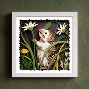 Op de afbeelding: Een vierkante witte lijst met een gelaagd papieren kunstwerk van een bruin-witte egel die tussen groen gras, hoge witte madeliefjes en kleine gele bloemetjes staat tegen een donkerbruine achtergrond met een geschulpte rand.