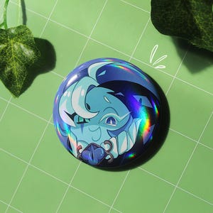 Op de afbeelding: Een ronde, blauwe knoop met een holografisch effect. De knoop toont een cartoonillustratie van een blauw wezen met wit haar en scherpe tanden. Het ontwerp heeft een fantasiethema.