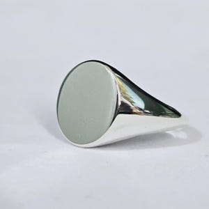 May include: A polished silver signet ring featuring a smooth, flat, circular face. The band is solid and reflective, showcasing a clean, metallic finish that catches the light. The design is simple and uniform, with a rounded top surface.