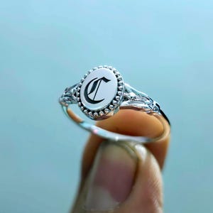 Peut inclure: Bague en argent avec une face ovale ornée de la lettre "C" en noir. La bague présente un motif de filigrane décoratif sur le corps et une bordure perlée autour de la lettre. La bague est tenue par les doigts d'une personne.